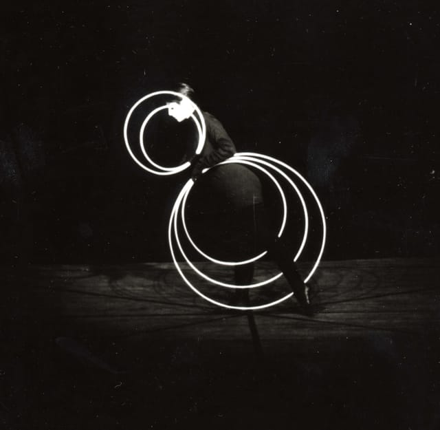 Hoop Dance performance. A dancer stands on one foot with her left foot stretched back, holding five white rings. Two rings form concentric circles near her head, three form concentric circles below her hips.